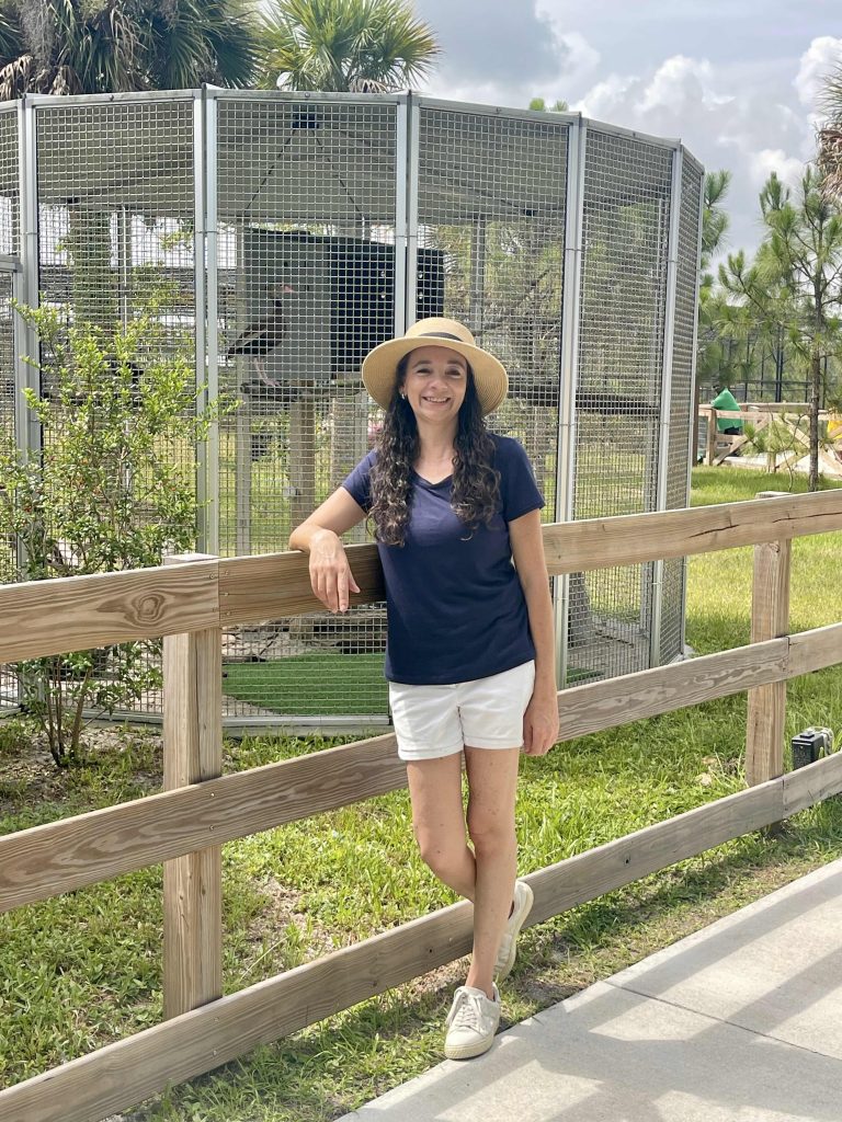 Woman in a sunhat standing and smiling by a wooden fence in front of a large bird enclosure at Busch Wildlife Sanctuary.